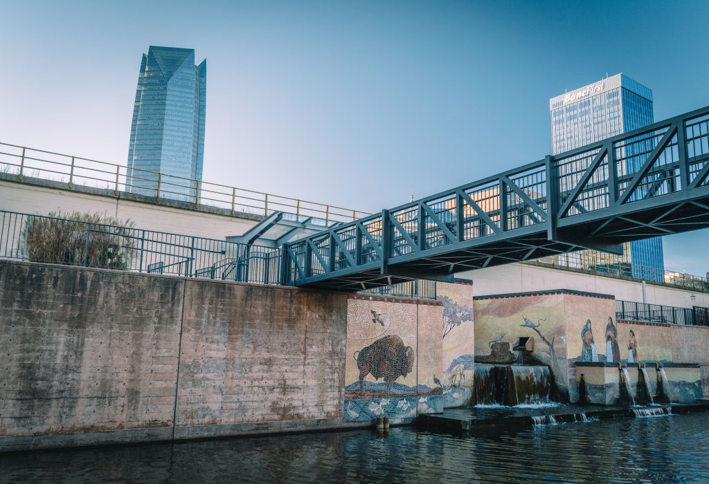 View of Oklahoma City's canal featuring a footbridge, murals, and skyscrapers in the background.